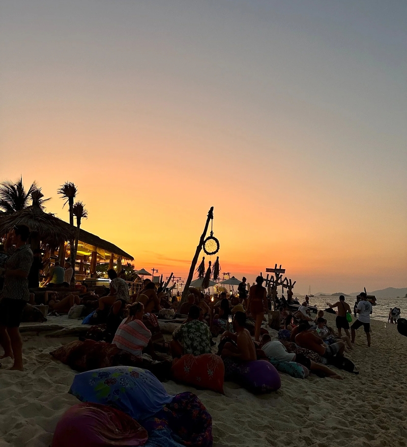 Sunset on Koh Lipe beach with vibrant beach bar, people relaxing on colorful cushions, and a stunning view of the horizon. Perfect spot for watching a fire show and enjoying the evening atmosphere.