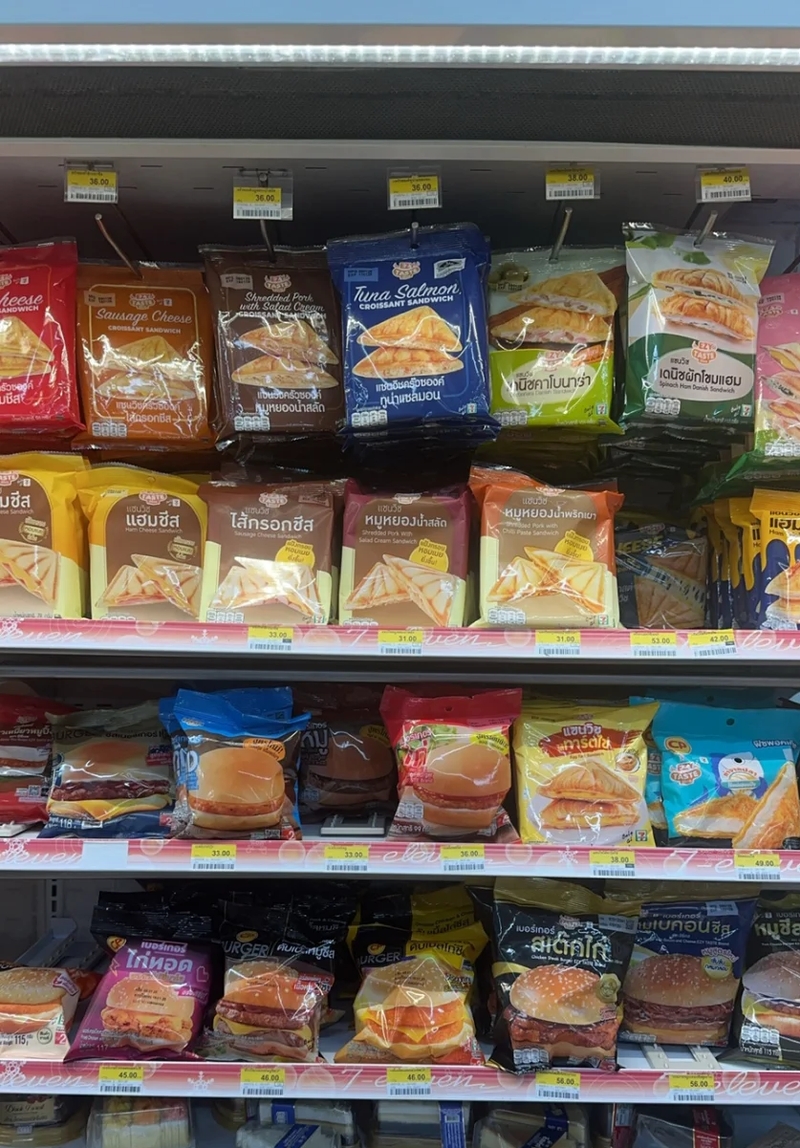 A well-stocked 7-Eleven shelf in Thailand displaying a variety of packaged sandwiches, croissants, and burgers. The packaging is colorful, with flavors like sausage cheese, tuna salmon croissant, and shredded pork. Price tags in Thai baht are visible above and below the products, making it easy for customers to choose their meal options.