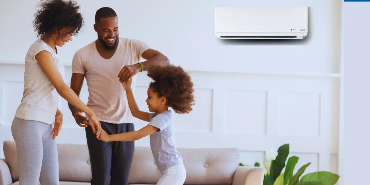 A happy family of three, consisting of a man, woman, and young girl, enjoying time together in a bright and comfortable living room. On the wall behind them, a white Trane Mitsubishi ductless mini split air conditioning unit is mounted, highlighting the room's modern climate control solution.