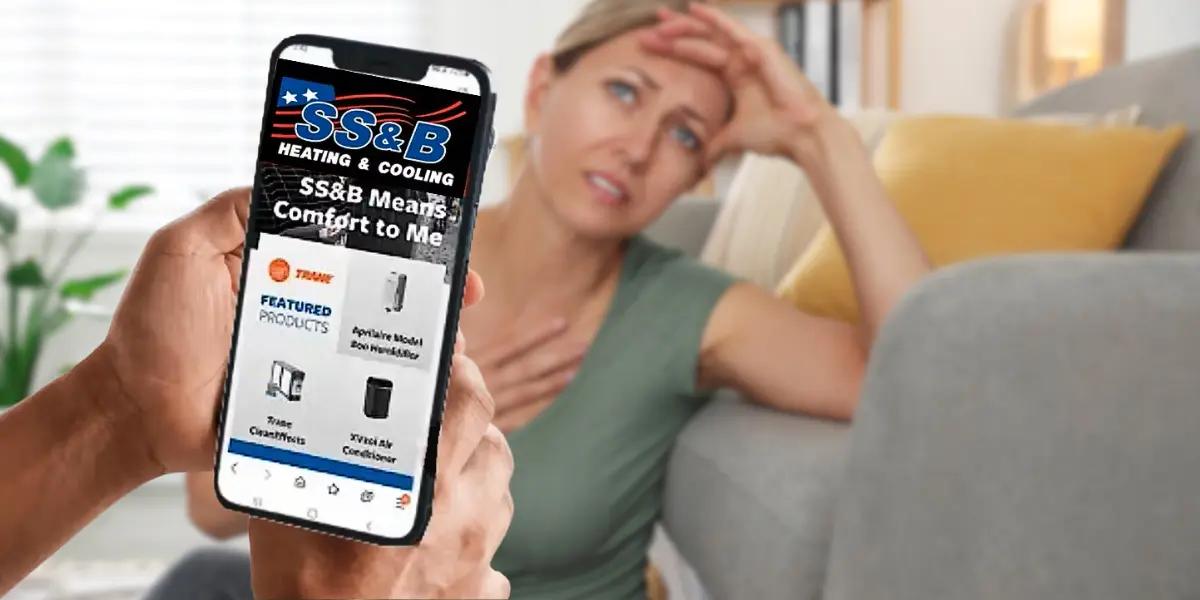 A woman looking distressed while sitting on a couch, wiping sweat from her forehead, indicating discomfort. In the foreground, a person holds a smartphone displaying the SS&B Heating & Cooling website, showcasing Trane HVAC products, emphasizing air conditioning solutions for home comfort.