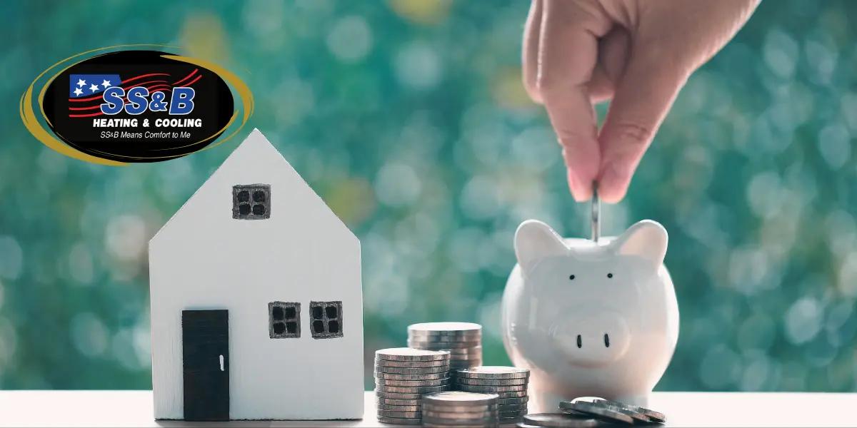 A small model house and stacked coins next to a hand placing a coin into a piggy bank, symbolizing energy savings and home efficiency with SS&B Heating & Cooling's logo prominently displayed.