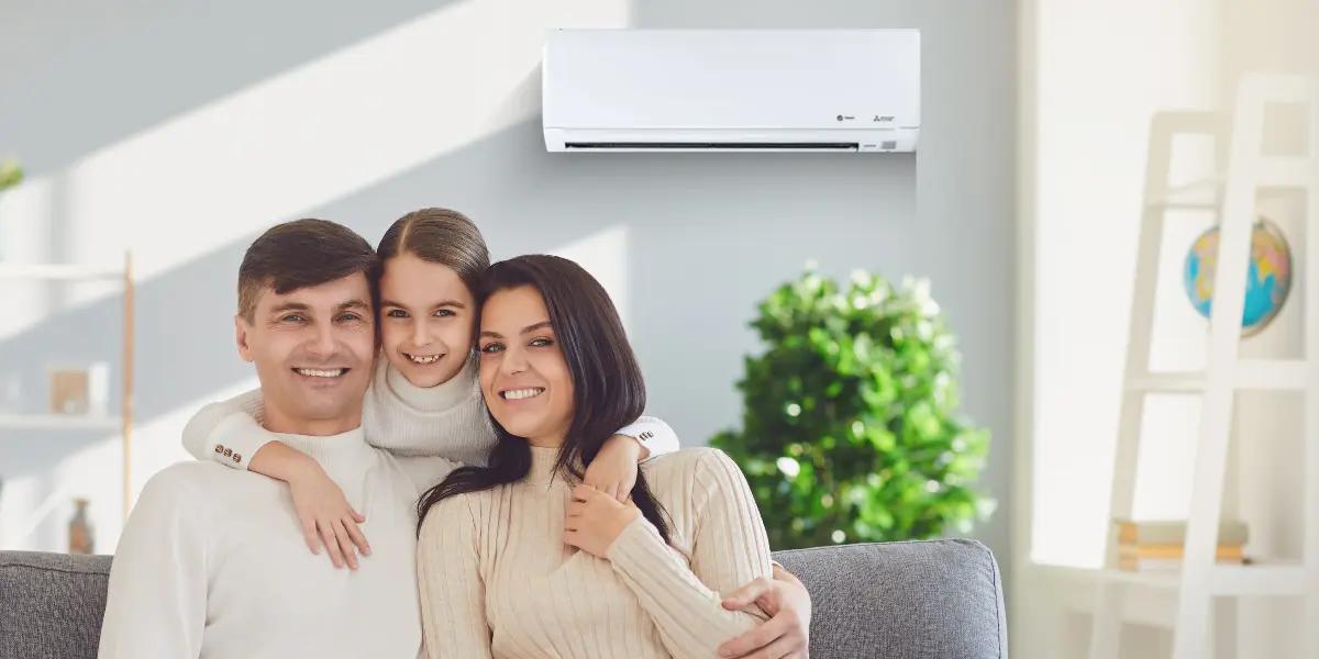 A smiling family of three, sitting on a couch in a cozy, sunlit living room with a green plant in the background. A white ductless Mitsubishi air conditioning unit is mounted on the wall above them, highlighting a comfortable and climate-controlled home environment.