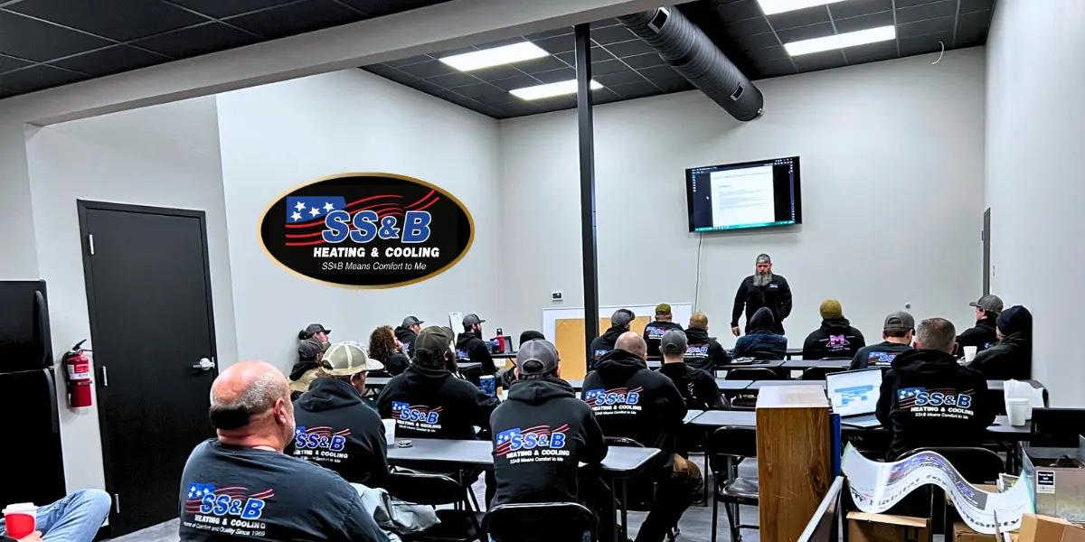 SS&B Heating & Cooling team meeting in a training room with technicians seated at tables and a trainer presenting on a screen, with the SS&B logo angled on the wall.