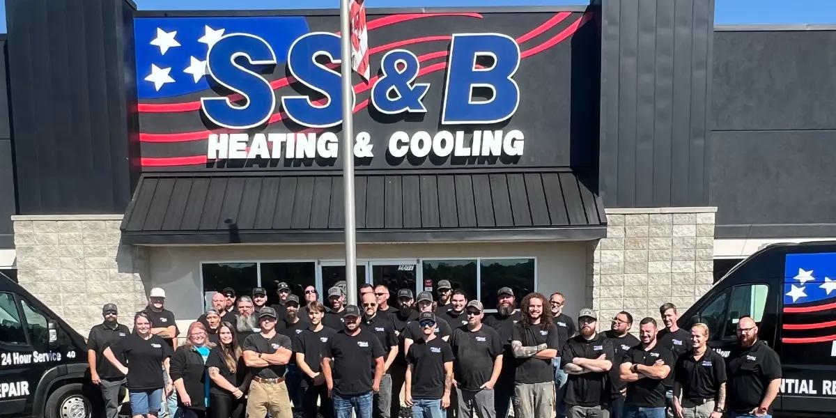 A group of employees from SS&B Heating & Cooling in Springfield, MO, standing in front of the company's building. The group, wearing black SS&B-branded shirts, is gathered under a large sign featuring the company name in bold letters with stars and stripes. Two company vans are visible on each side of the group, showcasing the company's services. The team appears unified and professional, representing SS&B Heating & Cooling in Springfield, MO.