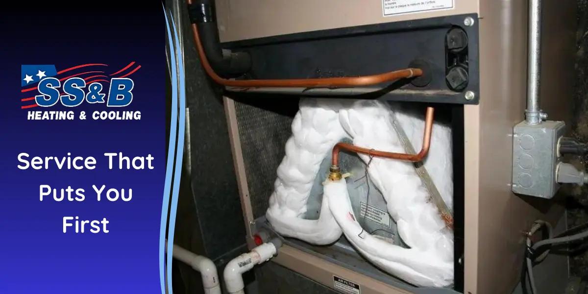 A frozen evaporator coil in an HVAC unit, covered in ice, indicating a common issue that restricts airflow. SS&B Heating & Cooling logo and "Service That Puts You First" slogan displayed on the left side.