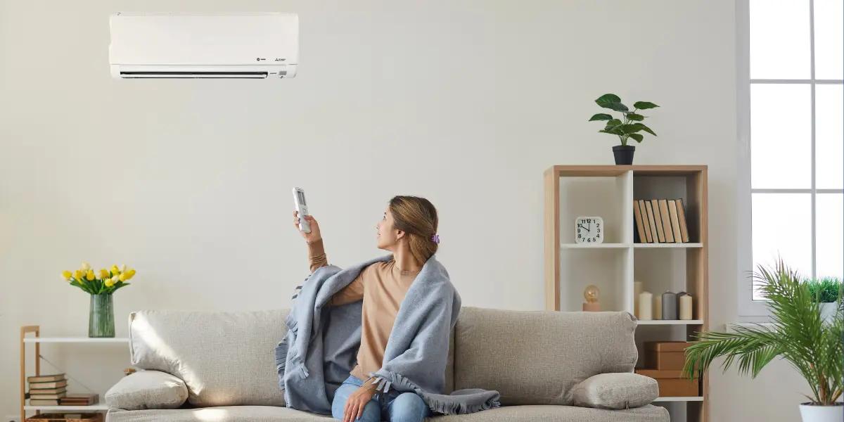 A woman sitting on a couch using a remote control to operate a wall-mounted ductless mini-split HVAC unit in her home.