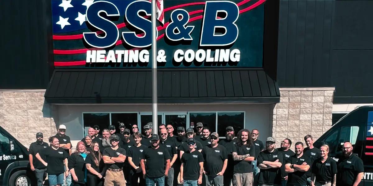 A group of employees from SS&B Heating & Cooling in Springfield, MO, standing in front of the company's building. The group, wearing black SS&B-branded shirts, is gathered under a large sign featuring the company name in bold letters with stars and stripes. Two company vans are visible on each side of the group, showcasing the company's services. The team appears unified and professional, representing SS&B Heating & Cooling in Springfield, MO.