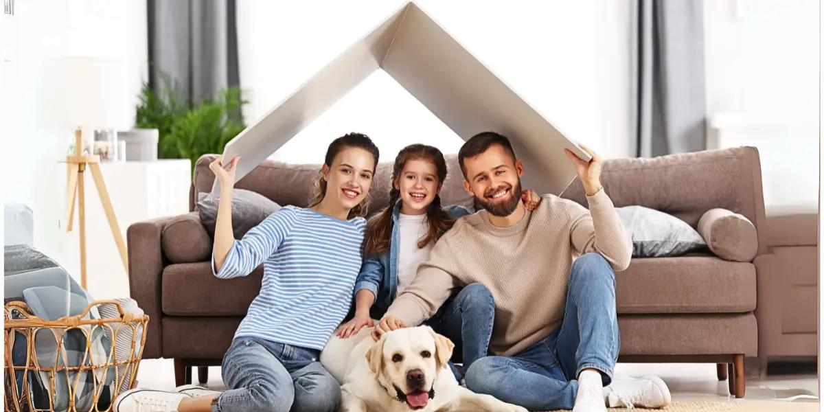 A happy family with a young child and their dog sitting together in a cozy living room, holding a roof-like structure symbolizing home comfort and protection.