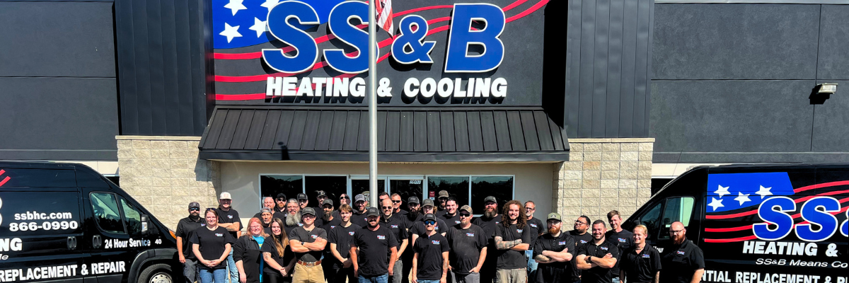 A group of employees from SS&B Heating & Cooling in Springfield, MO, standing in front of the company's building. The group, wearing black SS&B-branded shirts, is gathered under a large sign featuring the company name in bold letters with stars and stripes. Two company vans are visible on each side of the group, showcasing the company's services. The team appears unified and professional, representing SS&B Heating & Cooling in Springfield, MO.