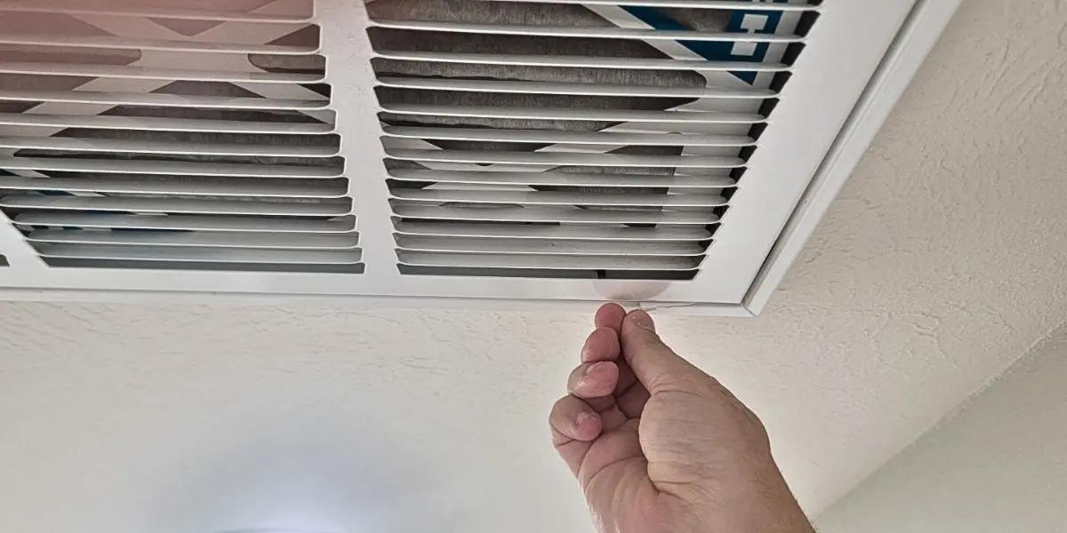 A hand reaching up to adjust an HVAC return air grille mounted on the ceiling, highlighting the importance of regular maintenance and cleaning for efficient airflow and HVAC system performance.