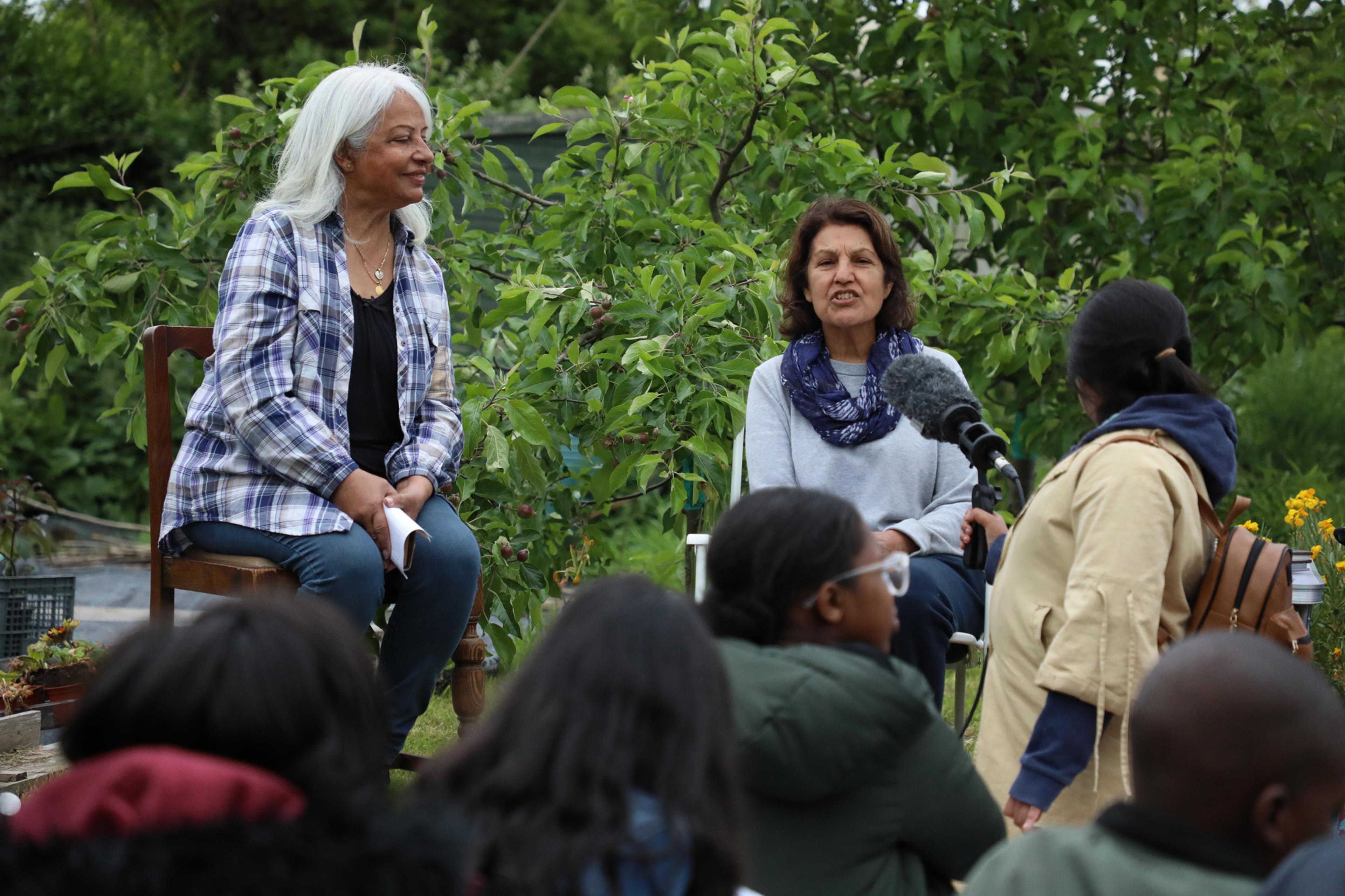 Two women site in chairs outdoors at an allotment, one speaking into a microphone held by a student