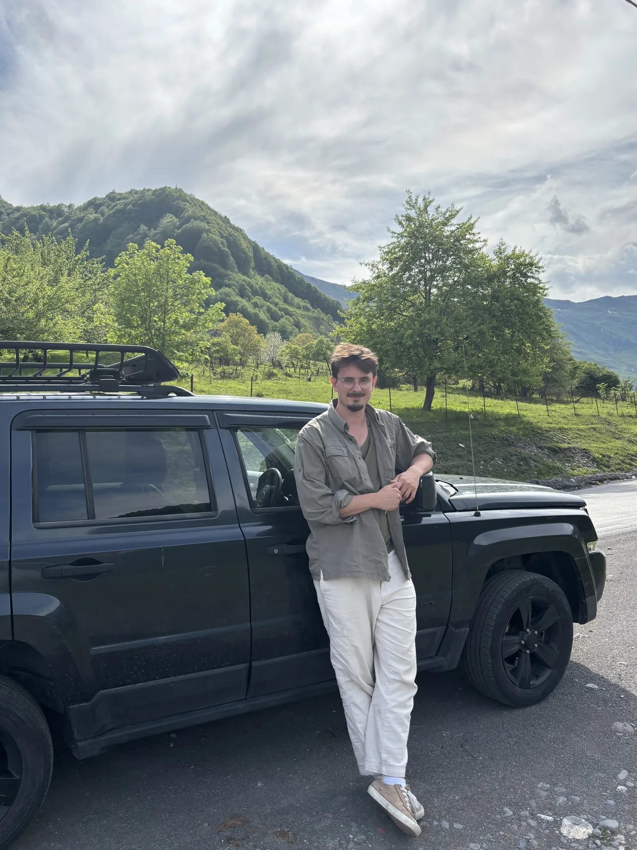Kevin mit Brille und hellem Hemd neben einem Geländewagen in grüner Berglandschaft