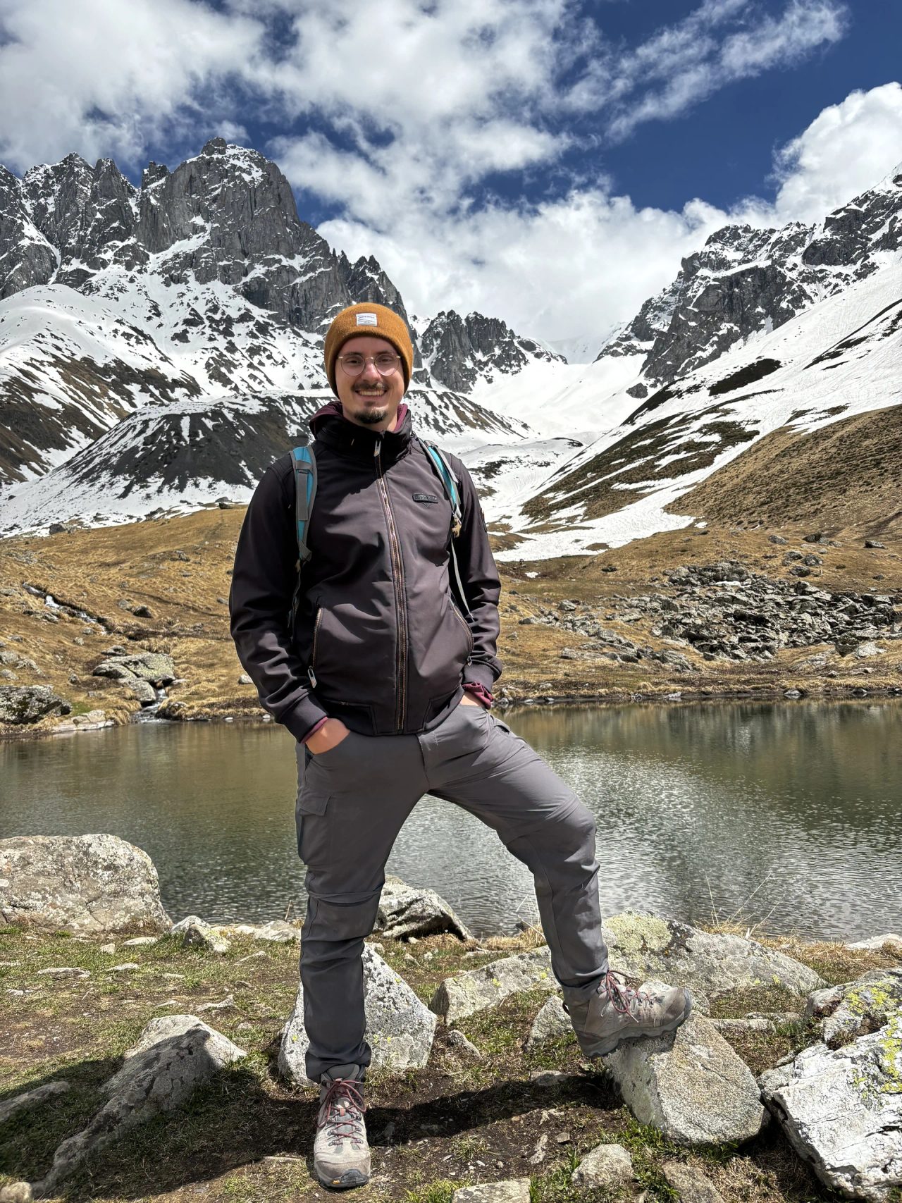 Kevin Otto Lais mit Rucksack an einem Bergsee vor schneebedeckten Gipfeln