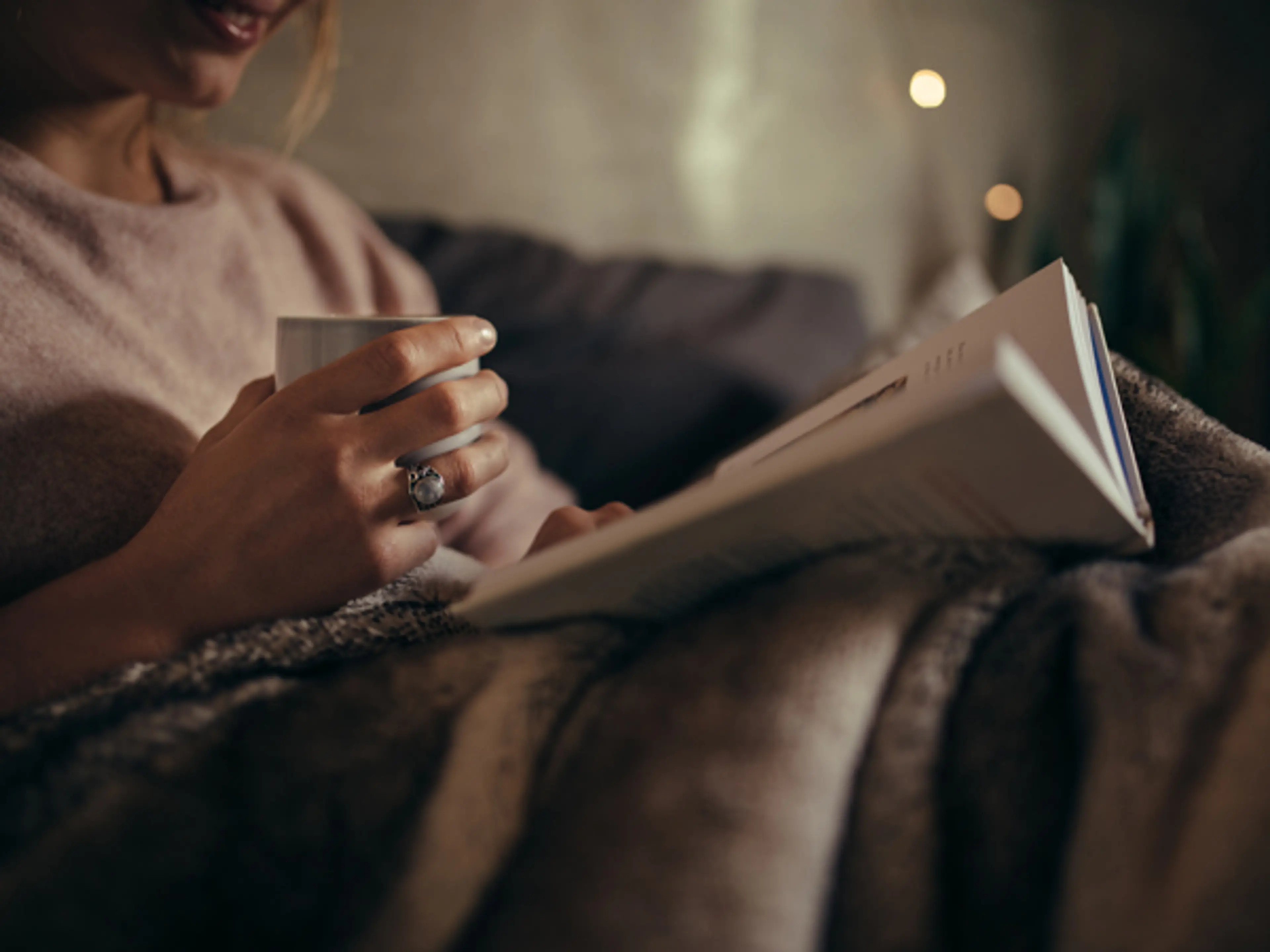 Dame sitter i sofa og leser bok med kaffekopp i hånda. Foto