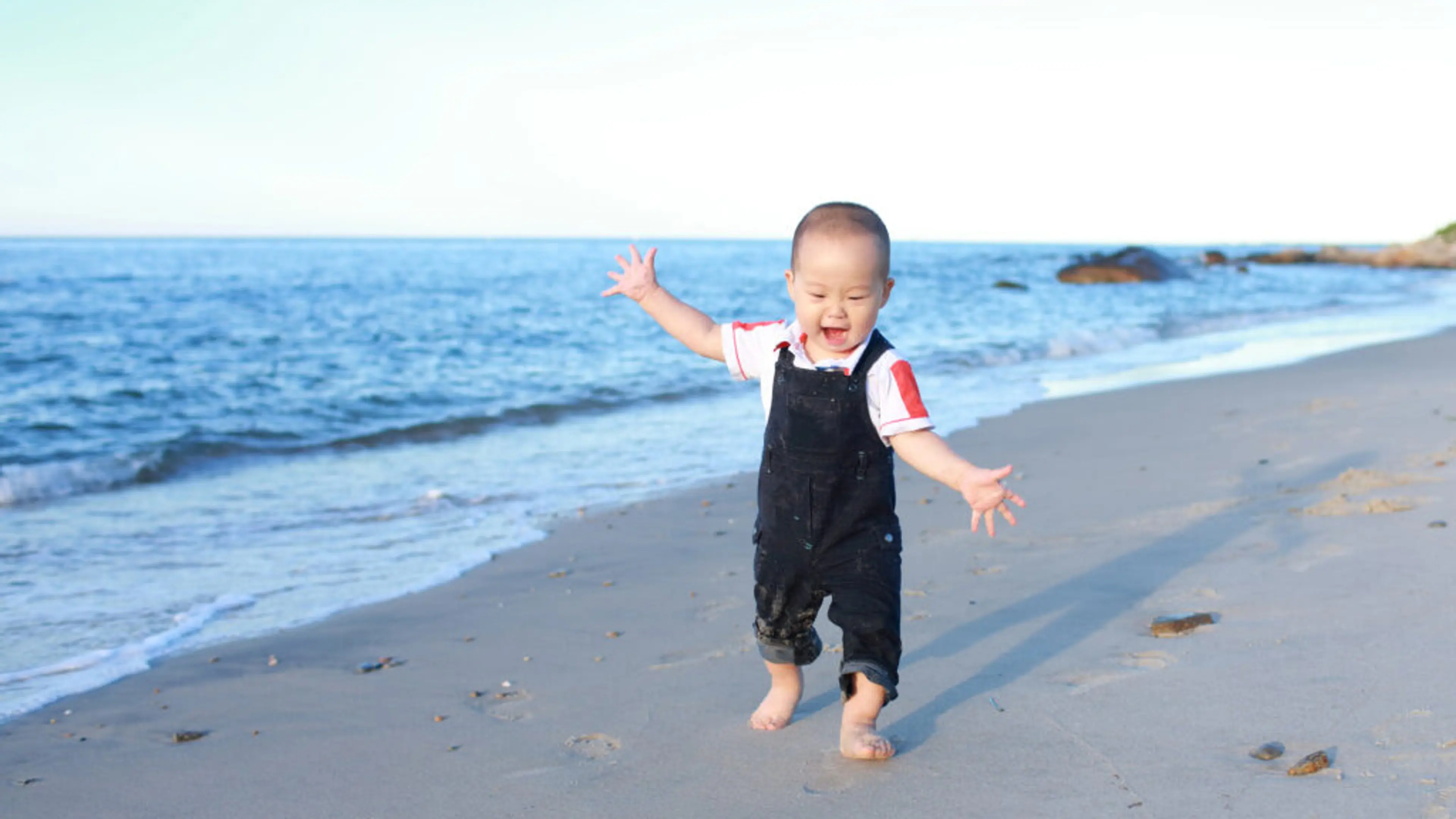Et barn som ser ut som det akkurat har lært å gå, går barbeint på en strand