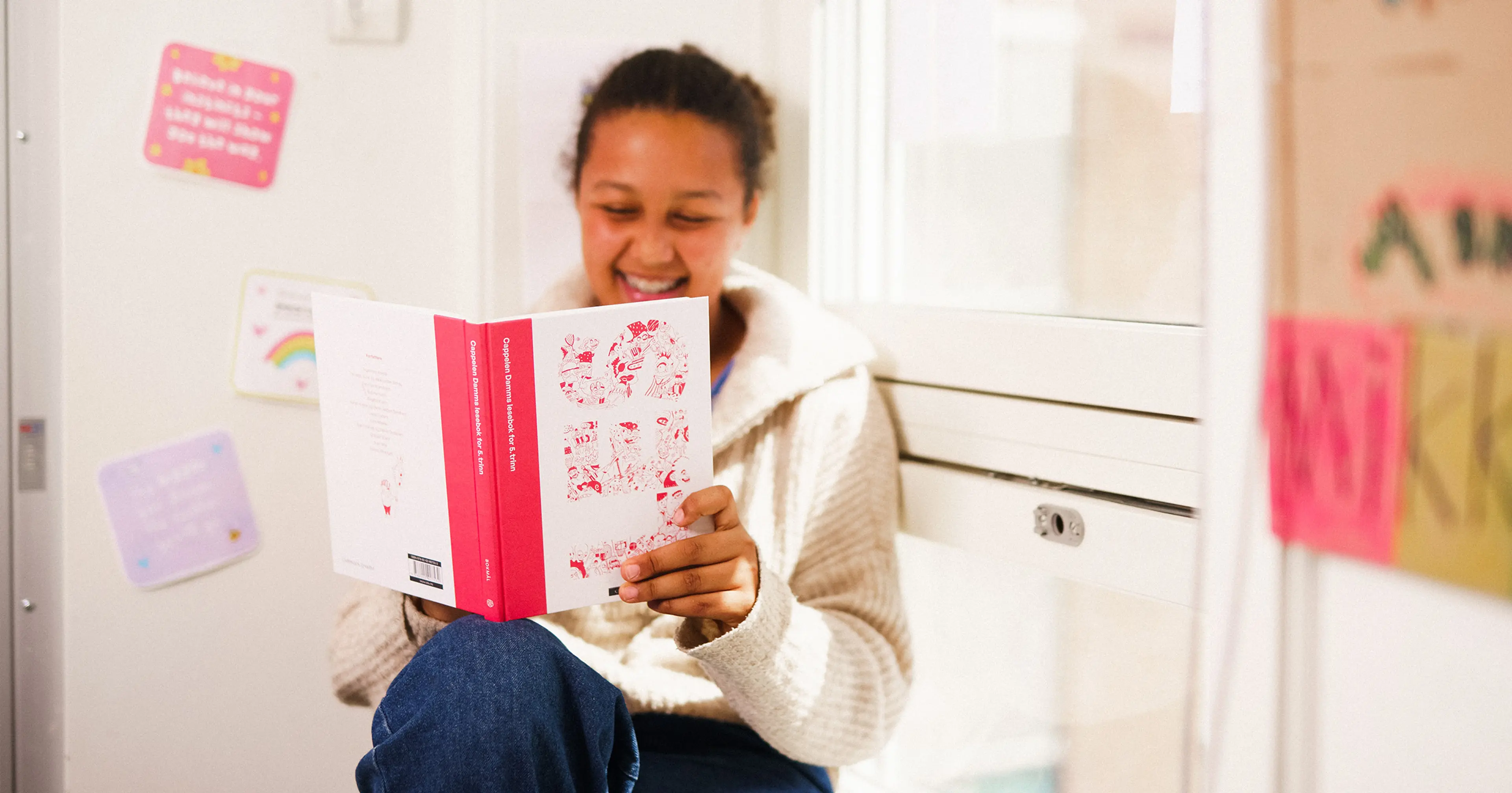 En smilende jente leser i leseboken LES fra Cappelen Damm i et klasserom.