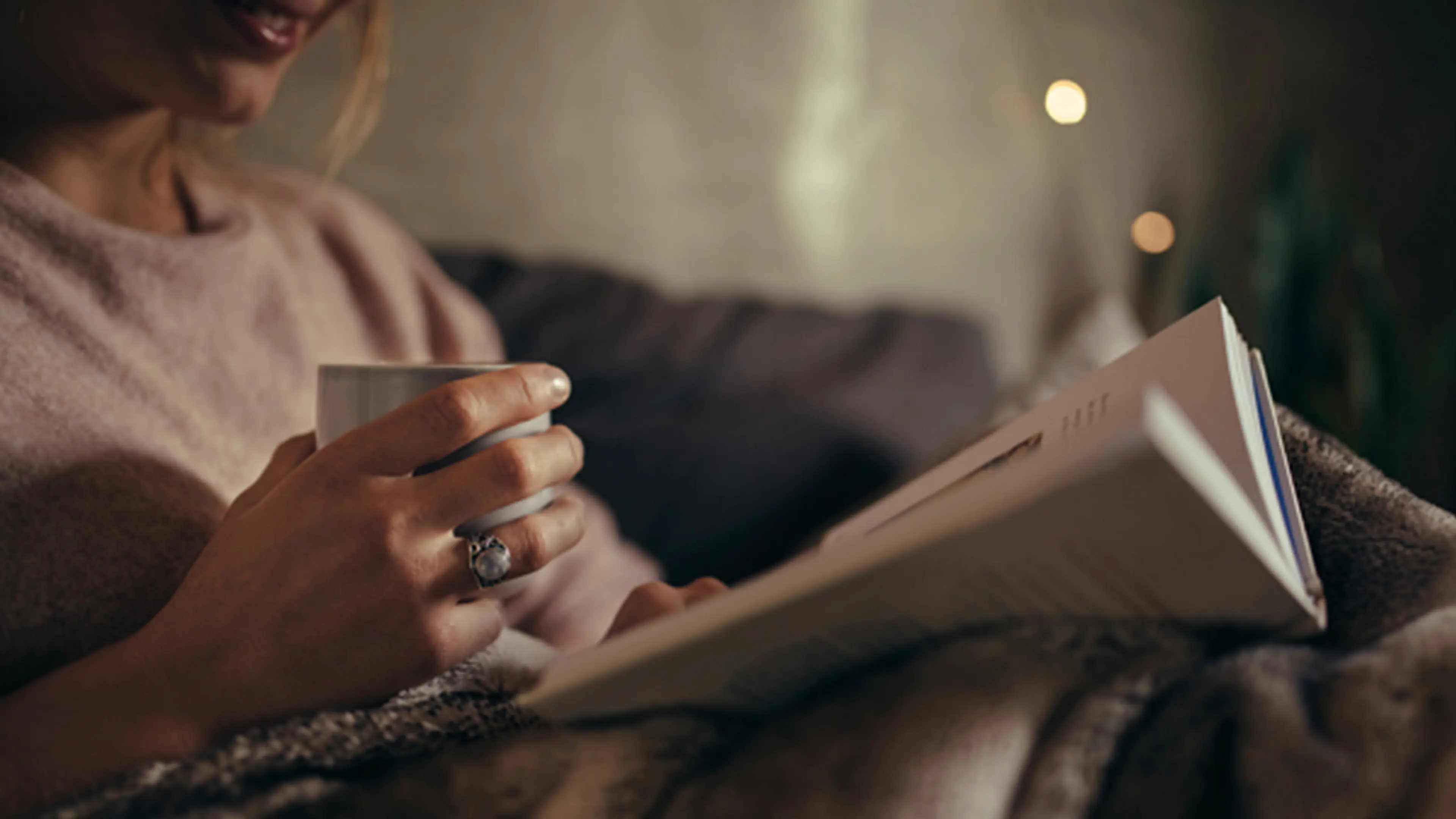 Dame som sitter i sofa og leser bok med kaffekopp i hånden