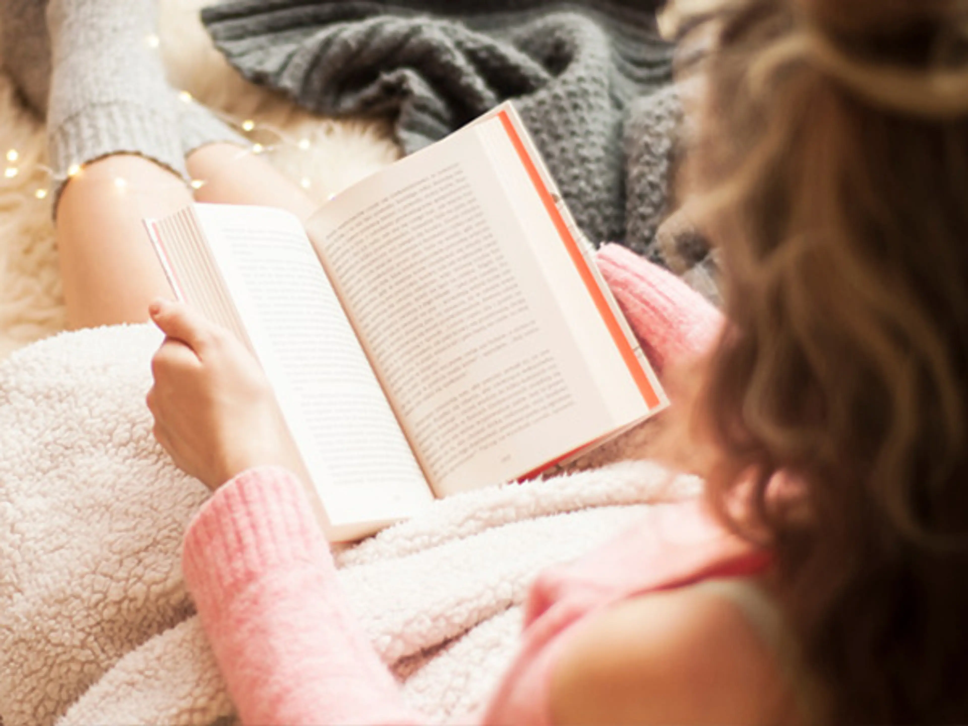 Dame i rosa genser som sitter og leser bok under et teppe. Foto
