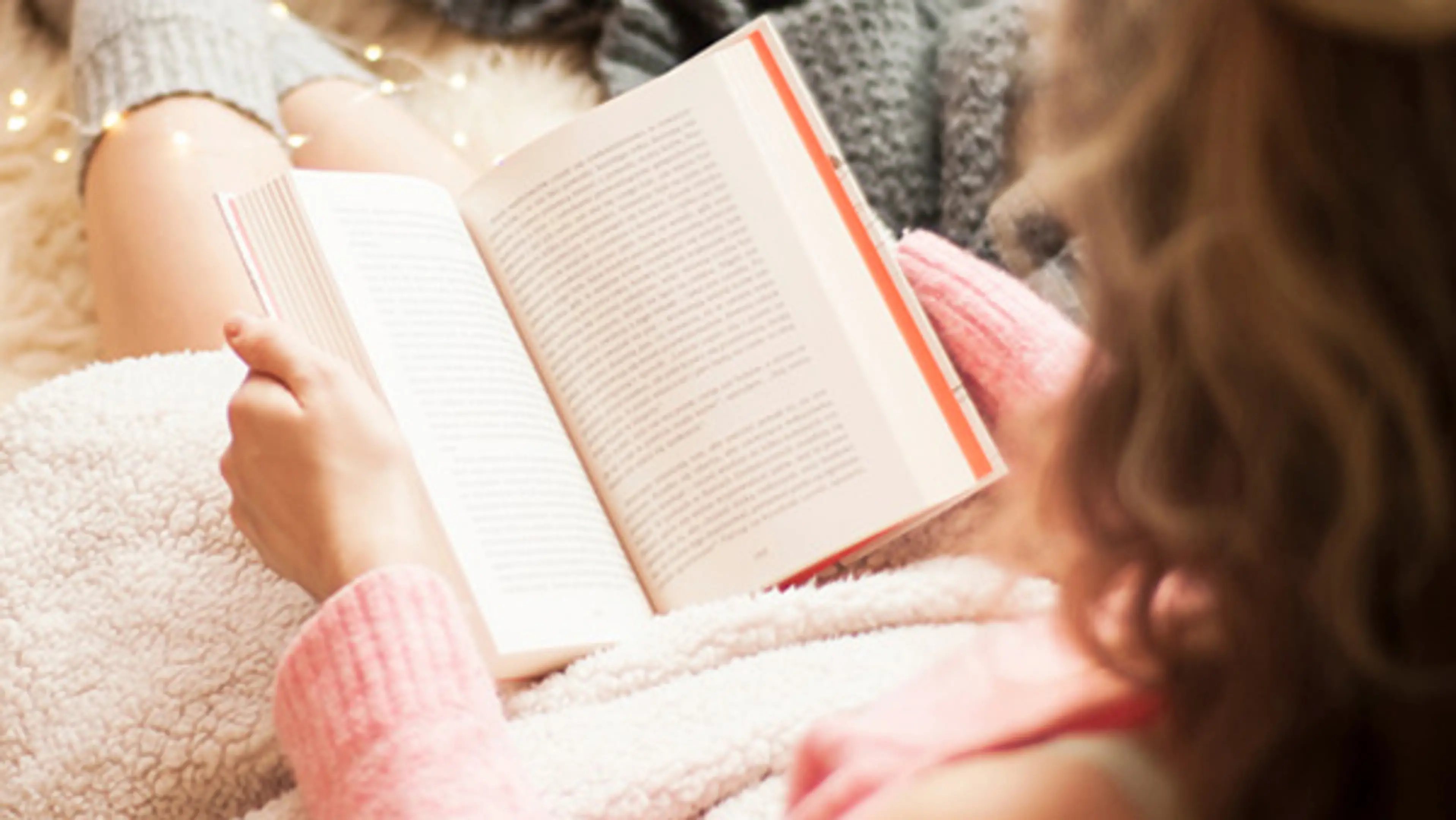 Dame i rosa genser som sitter og leser bok under et teppe. Foto