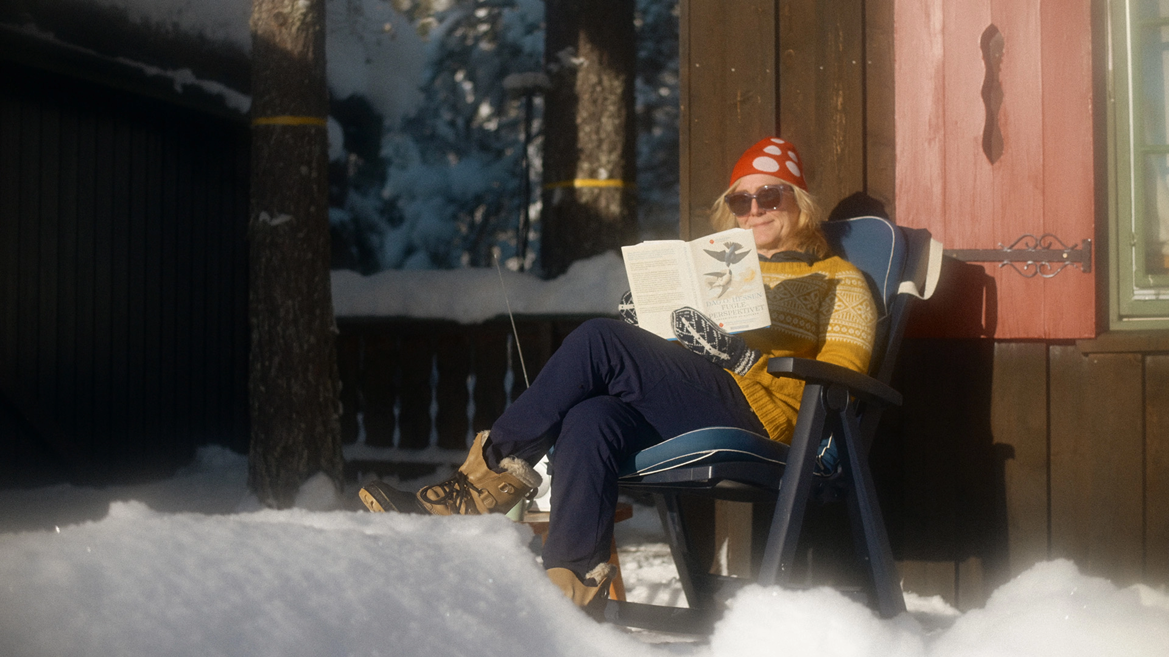 En kvinne med solbriller og gul strikkegenser sitter i en stol i hytteveggen og leser boka "Fugleperspektivet"