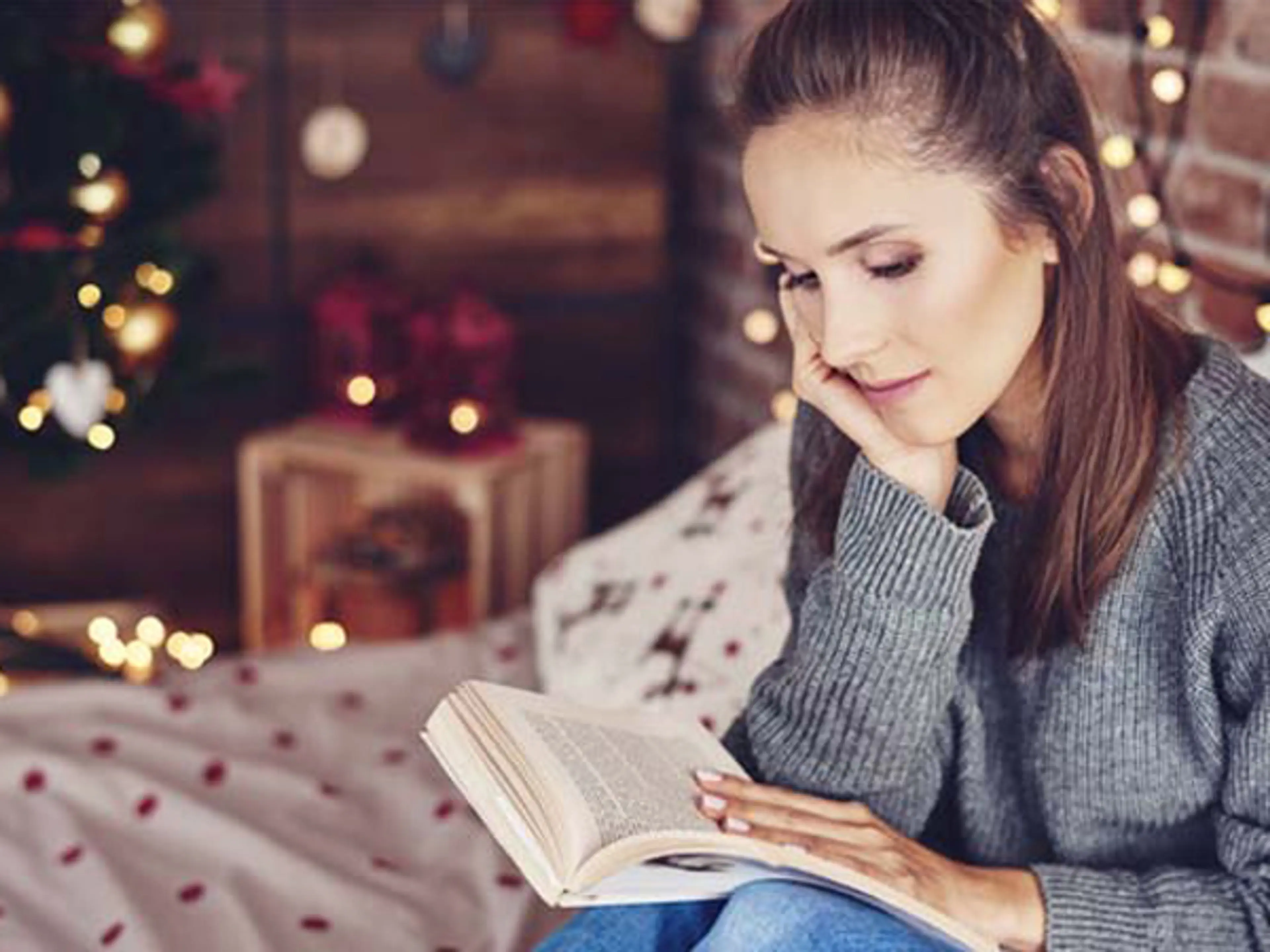 Dame som sitter og leser bok med julelys i bakgrunnen. Foto