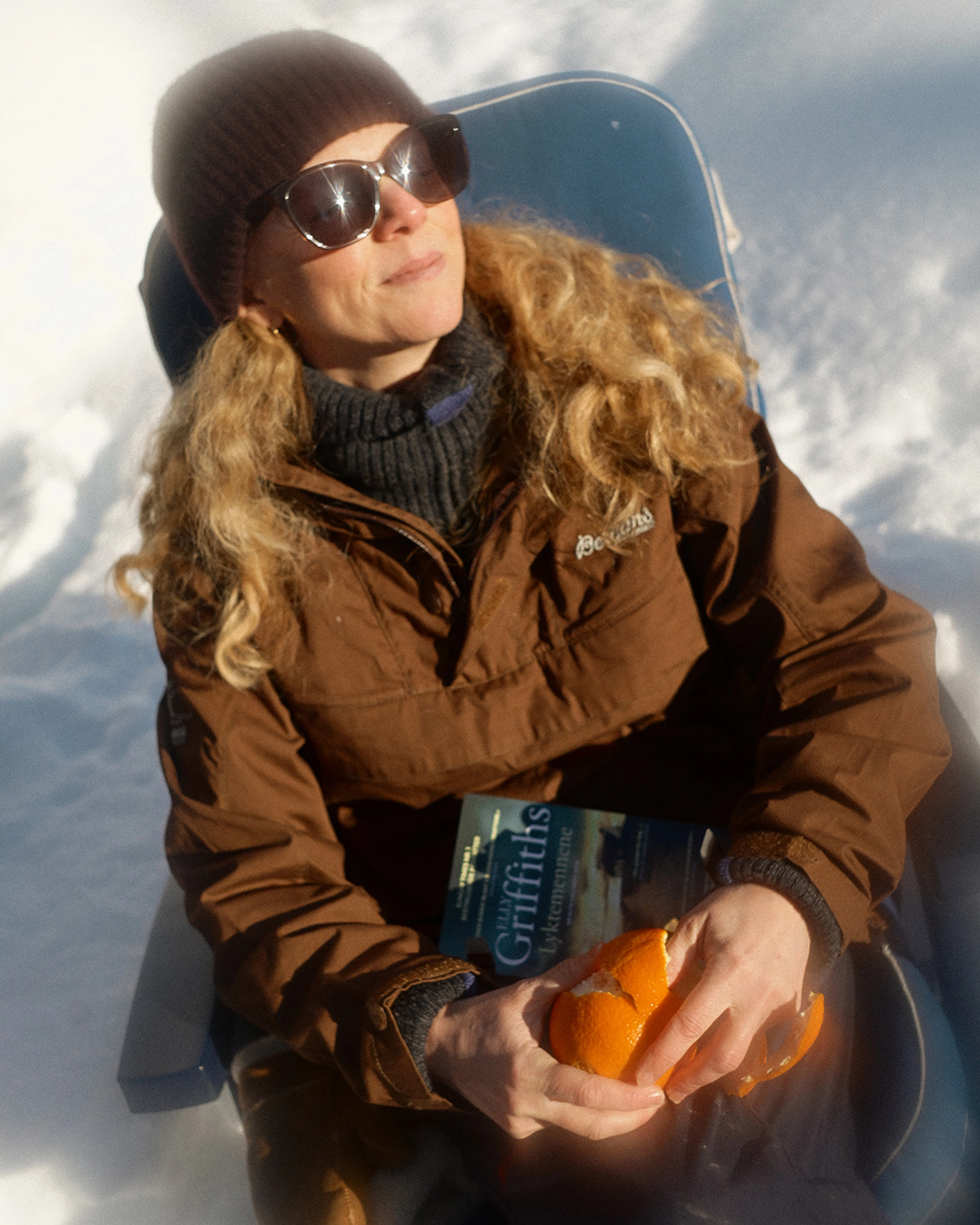 En kvinne med solbriller og ansiktet vendt mot solen sitter i en stol i snøen med en krimbok i fanget mens hun skreller en appelsin.