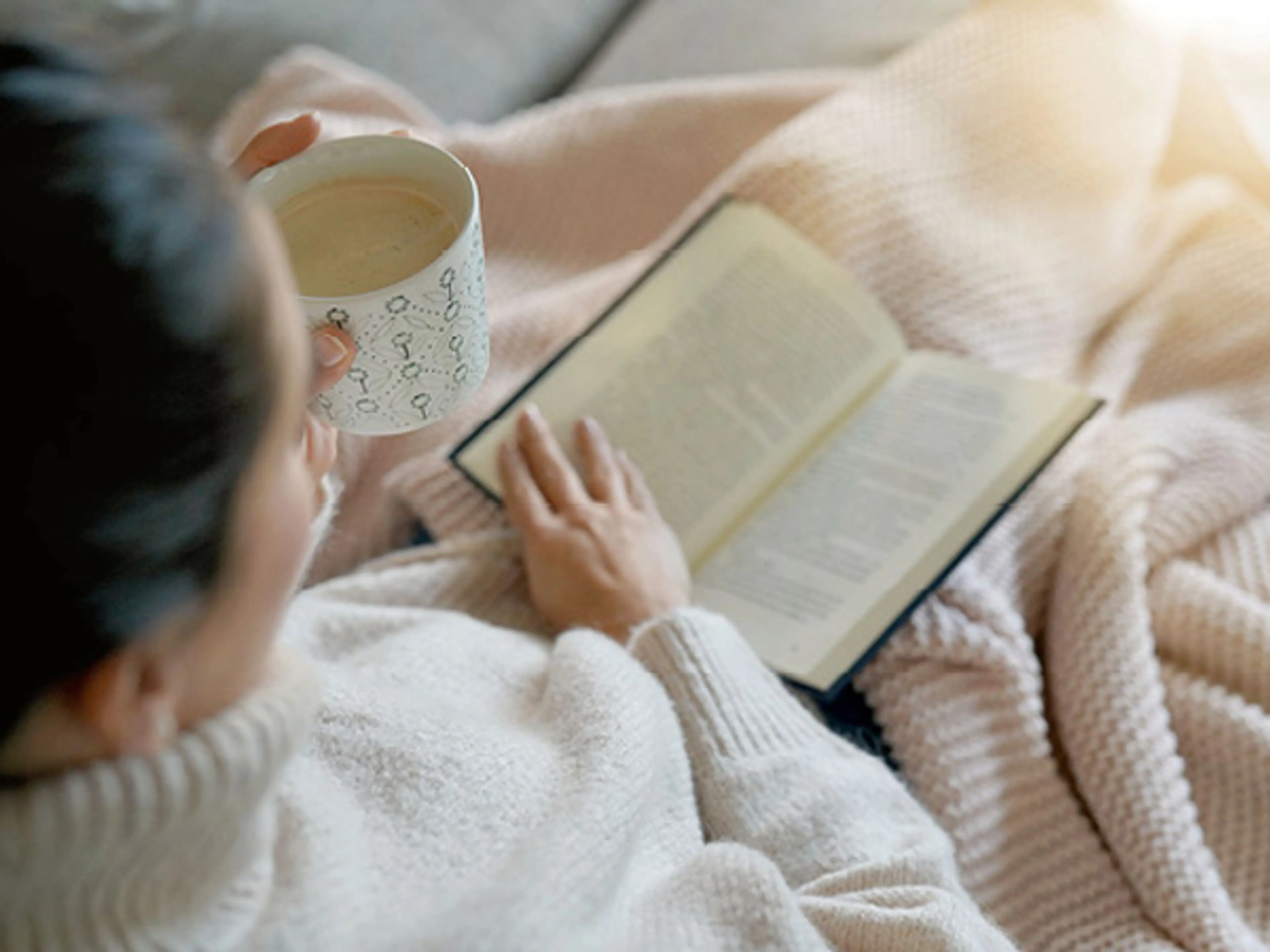 Dame i rosa genser som sitter og leser bok under et teppe. Foto