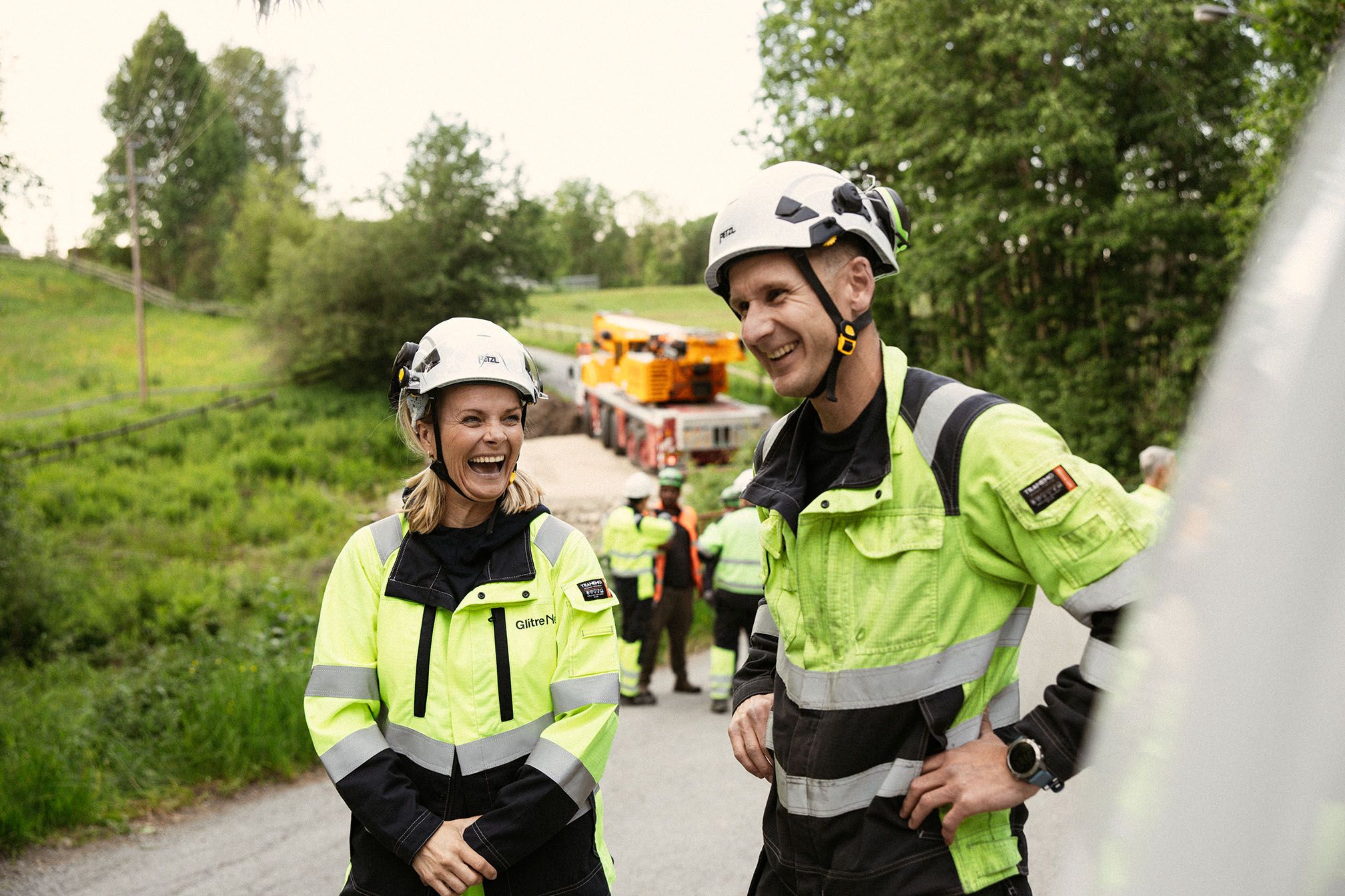 Dame og mann i arbeidstøy og hjelm. Ute. Glade.
