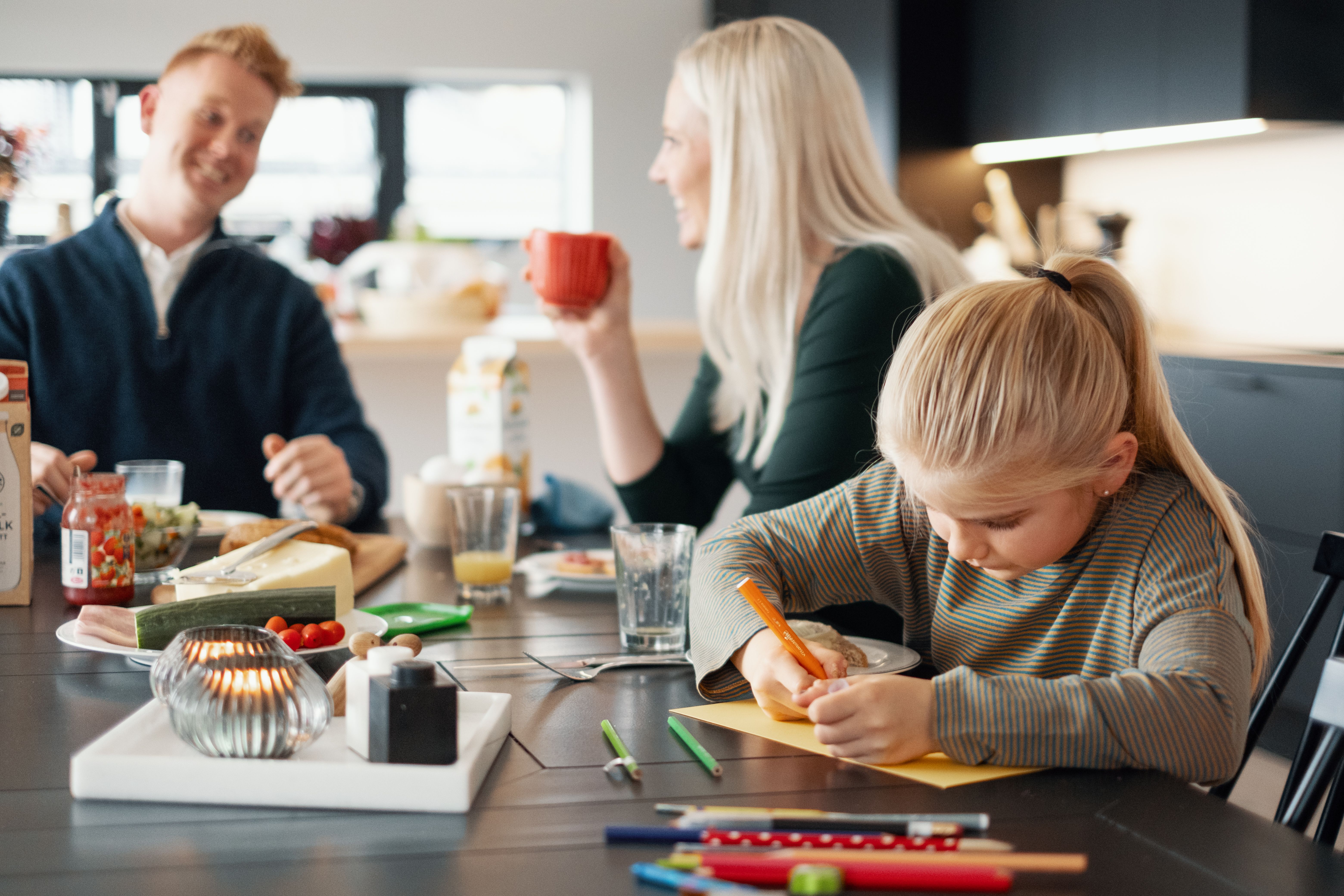 Familie ved kjøkkenbord. Mor og far drikker kaffe. Datter som tegner.