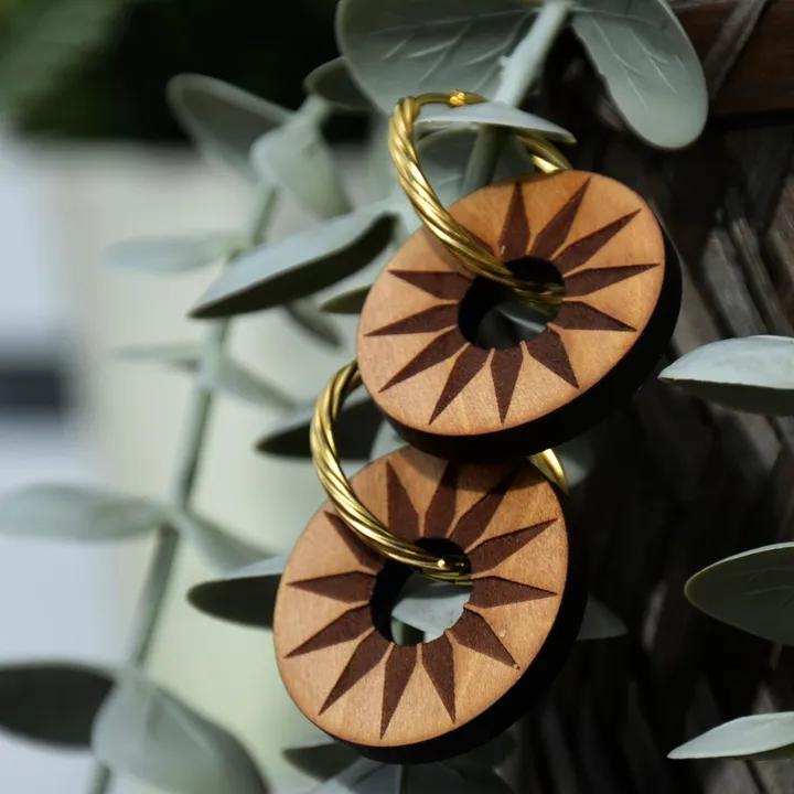 Pendentifs ronds en bois gravé d’étoiles, sur des créoles dorées, devant des feuilles d’eucalyptus
