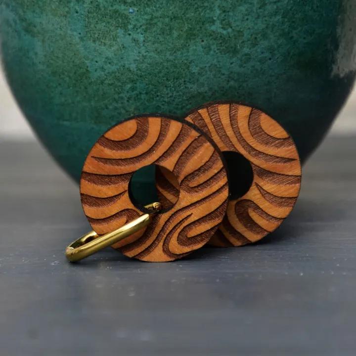 Pendentifs ronds en bois gravé de motifs ondulés, sur des anneaux dorés, devant un vase turquoise