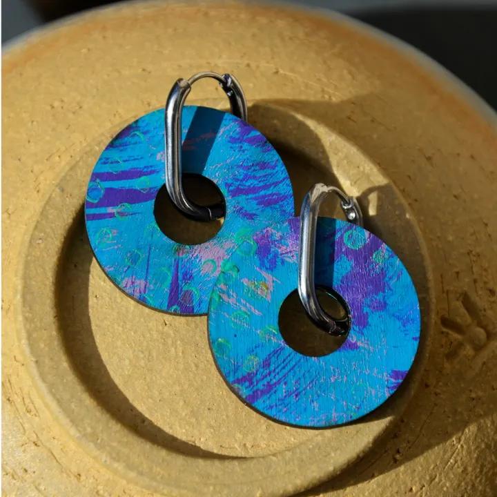 Pendentifs ronds peints en bleu et violet, sur des créoles argentées, posés sur une assiette en grès