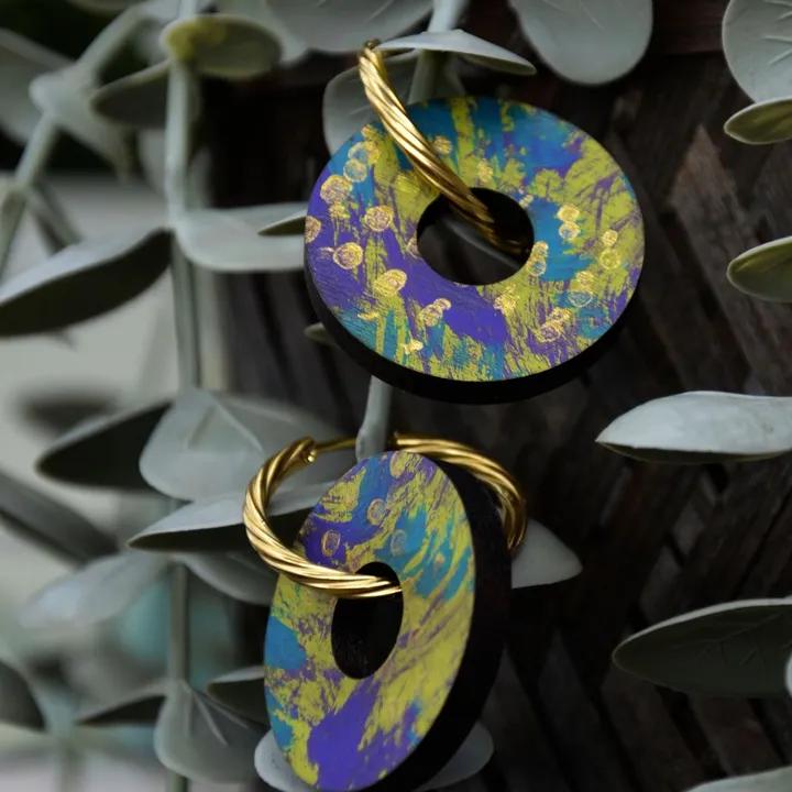 Pendentifs ronds peints en violet, jaune et turquoise, sur des créoles dorées, devant des feuilles d’eucalyptus