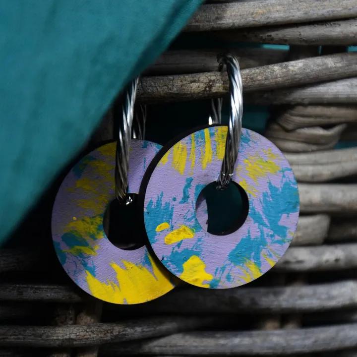 Pendentifs ronds peints en lilas, jaune et turquoise, sur des créoles argentées torsadées, devant un panier en osier