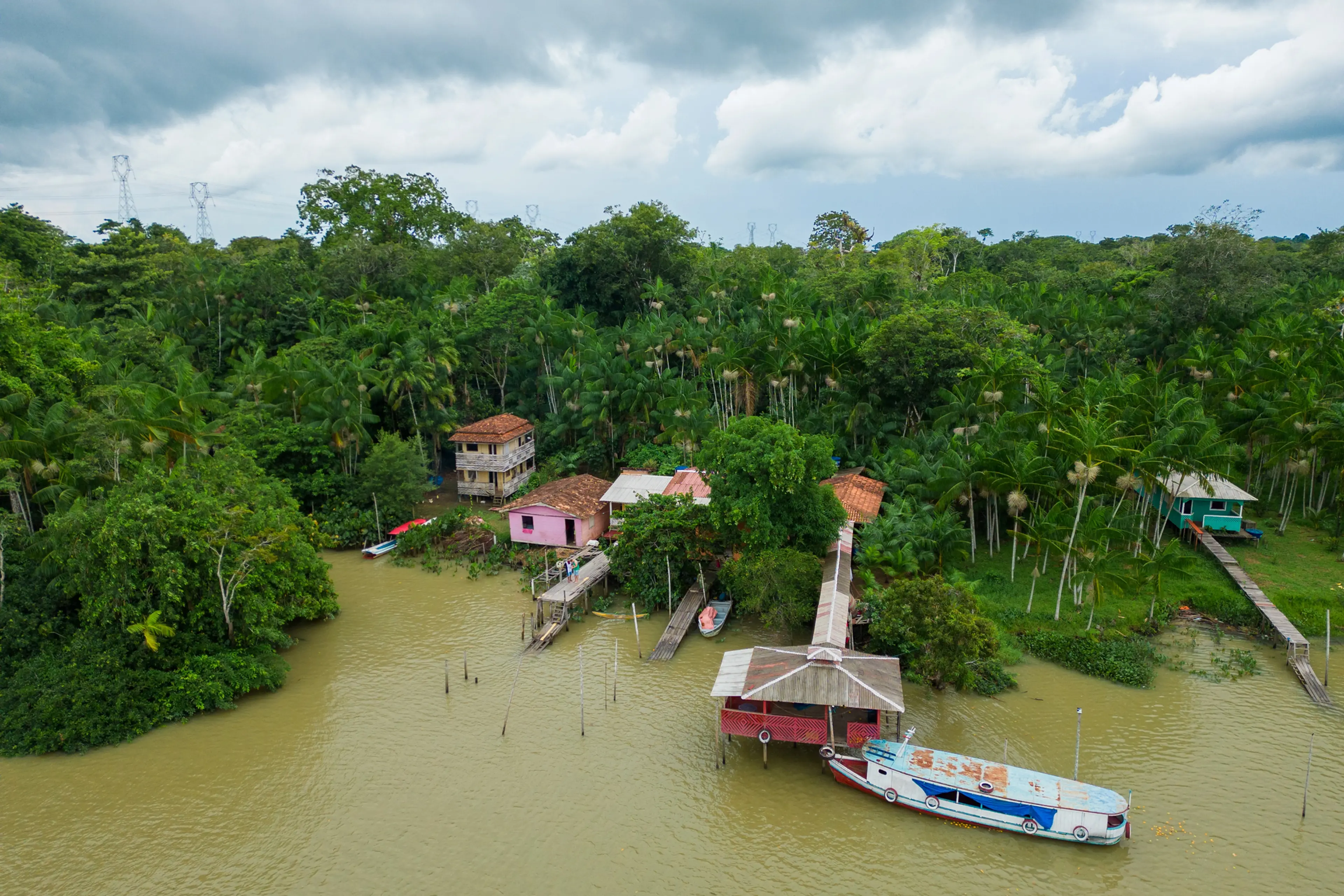 Aerial view of a small village on a muddy river, with colorful houses, wooden docks, and a boat, surrounded by dense jungle.