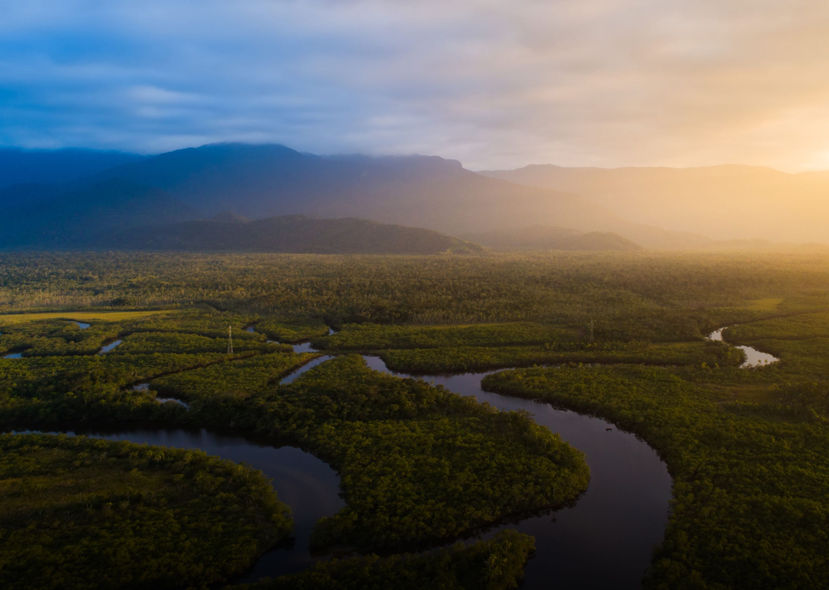 Aerial view of a winding river through a dense forest, with mountains and a split blue and golden sky.