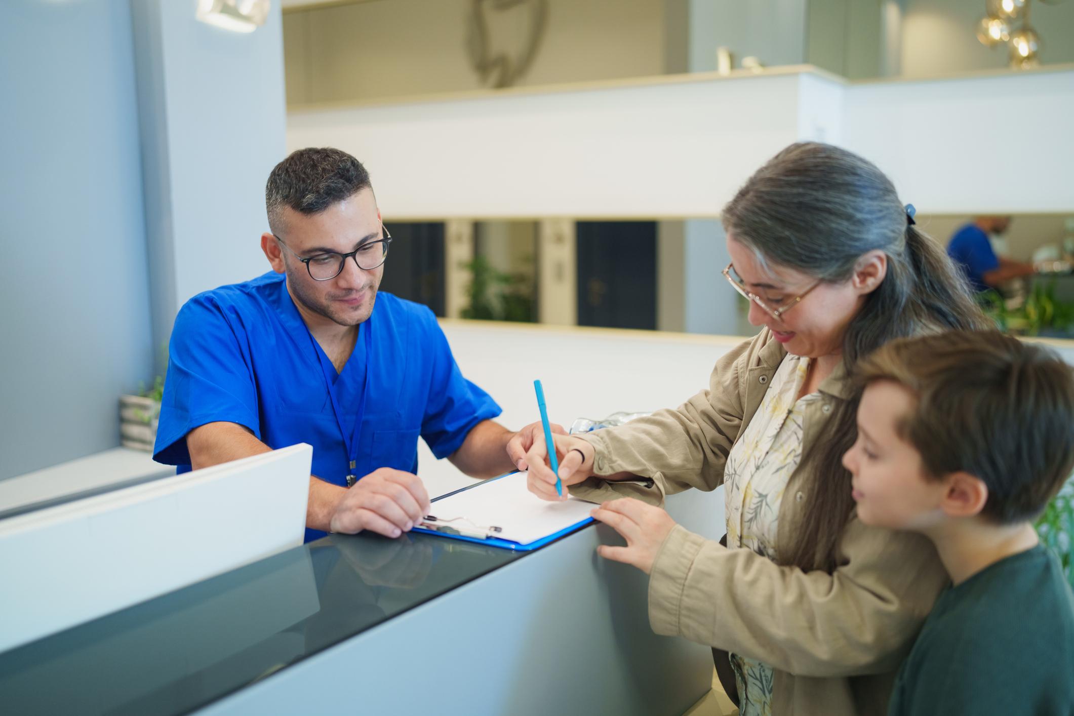 A friendly dentist in blue scrubs taking notes on a clipboard while discussing treatment details with a mother and her young son at the reception area of a modern dental clinic