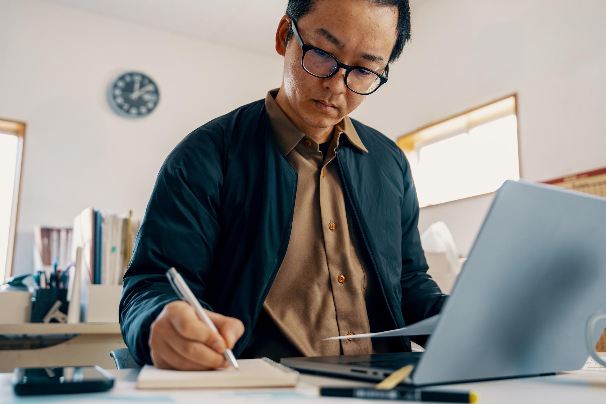 Man writing notes while working on a laptop in an office setting.