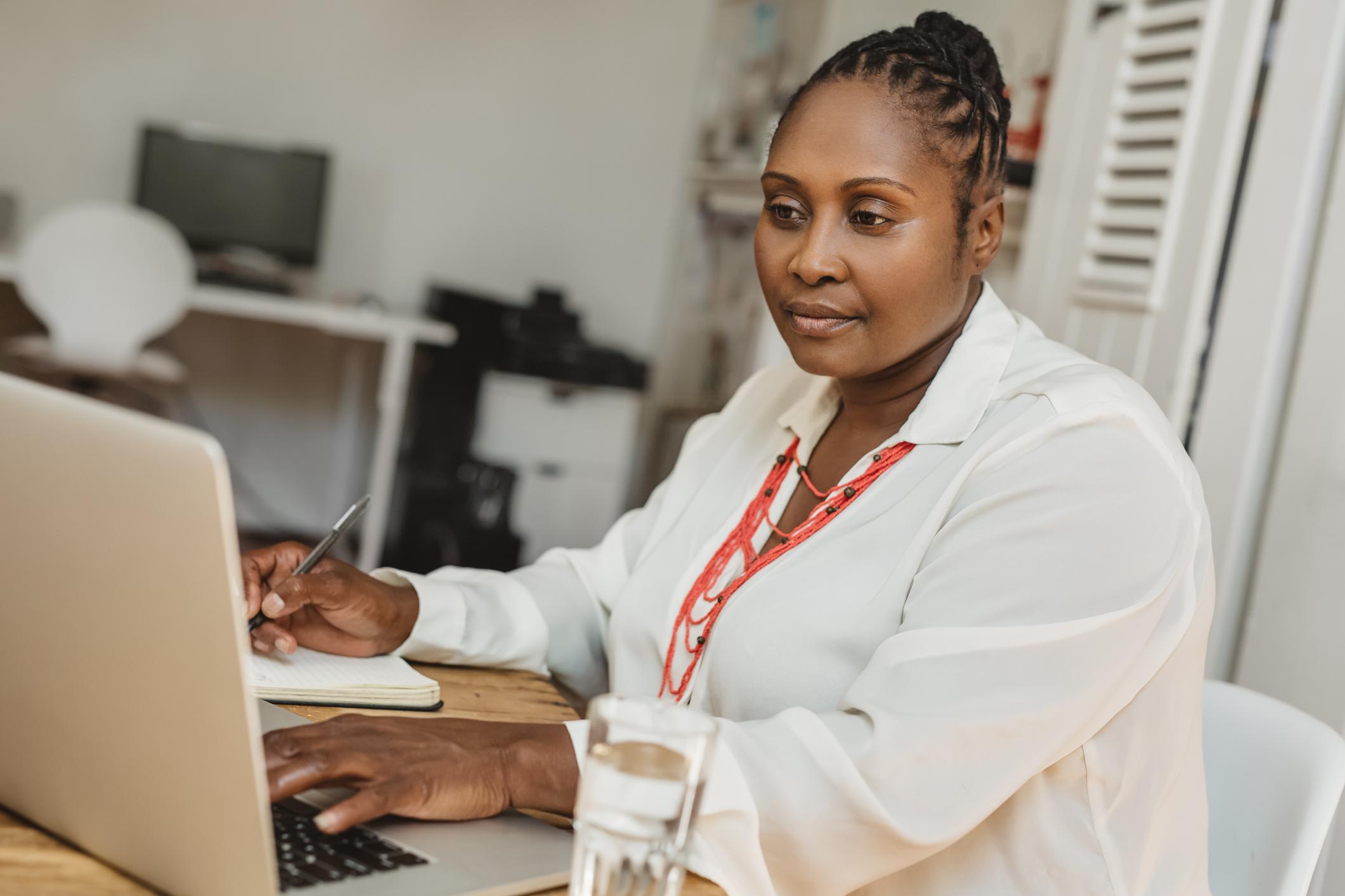 Woman working on a laptop and taking notes in an office setting.