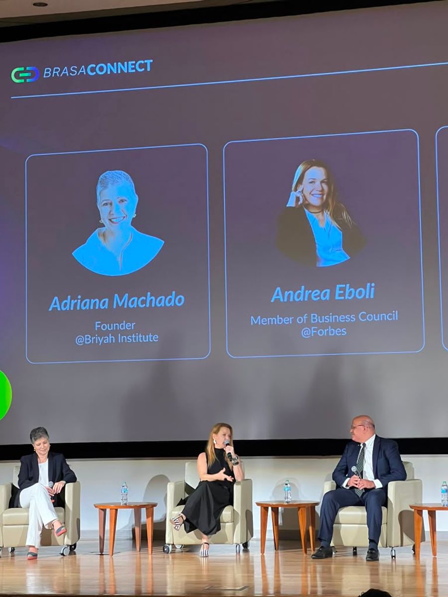 Andrea Eboli no palco de um painel do BrasaConnect; o telão apresenta os participantes