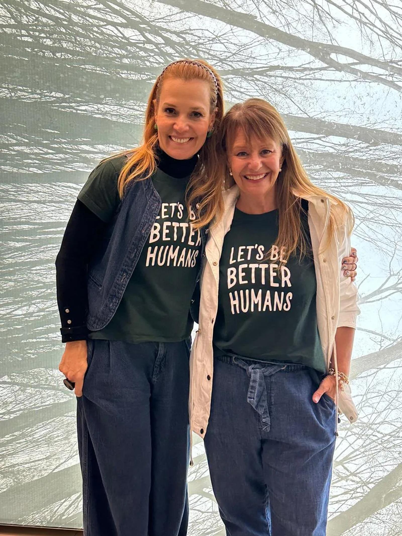 Duas participantes da Confraria Let’s Be posam juntas diante do painel de árvores, de camiseta Let’s Be Better Humans