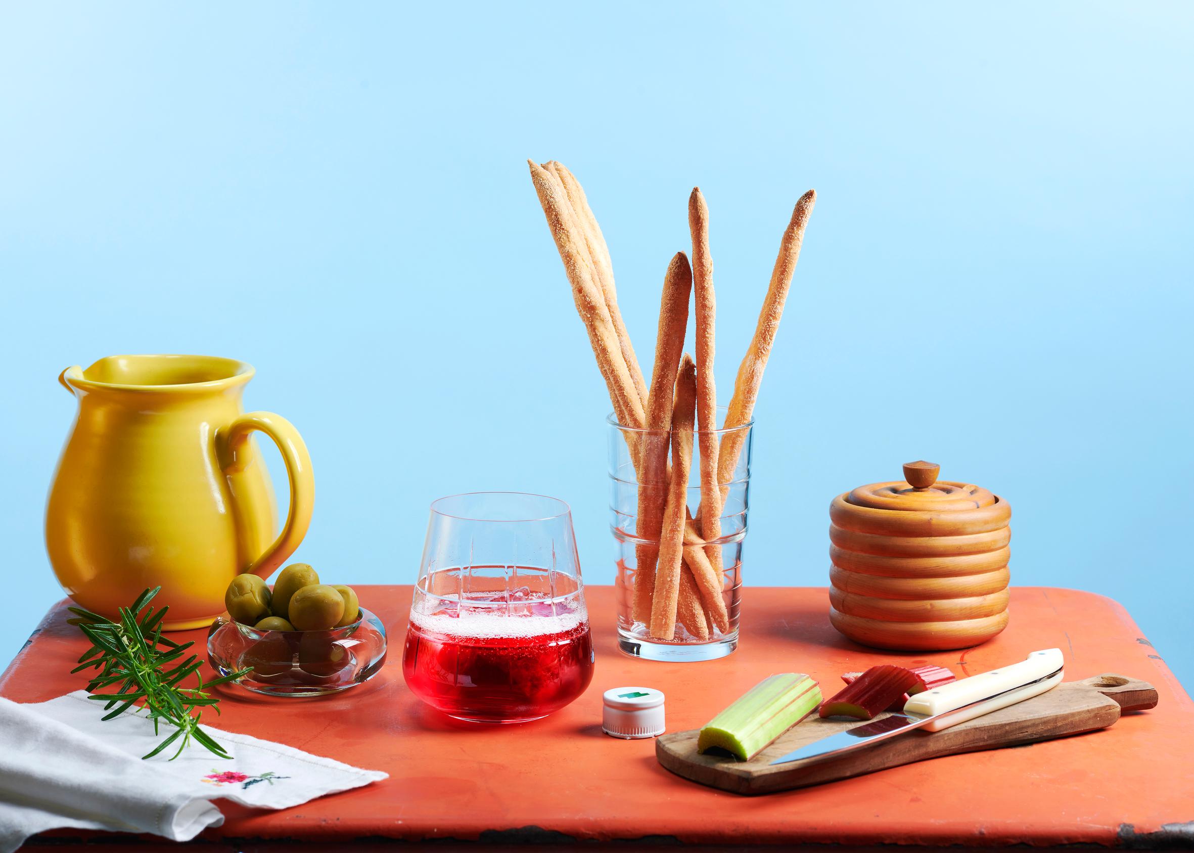 Scene with a yellow ceramic jug, olives, a sprig of rosemary, breadsticks, and fresh rhubarb