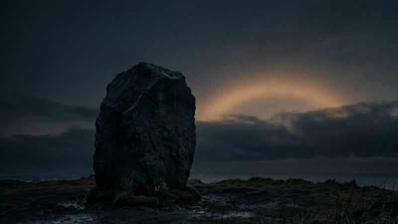 A large weathered standing stone sits alone on a barren landscape beneath a dark,
