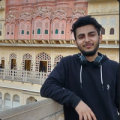 A young man in a black hoodie with headphones around his neck stands leaning on a railing, posing in front of an ornate pink and yellow historic building with intricate windows and architectural details.