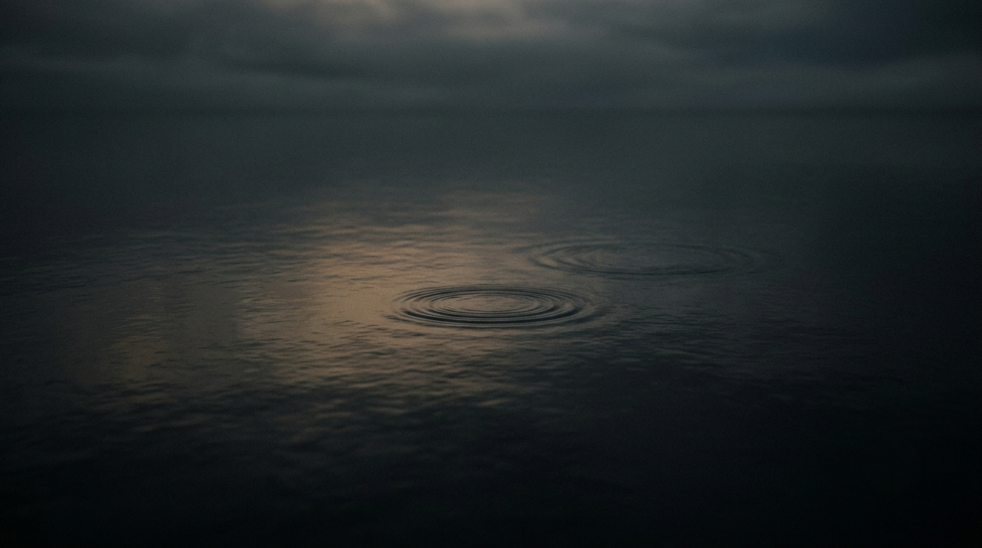 A dark, cinematic editorial photograph of faint concentric ripples expanding across a quiet, shadowed body of water under an overcast twilight sky.