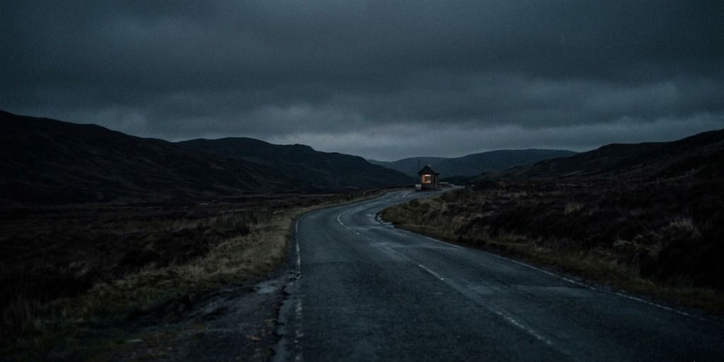 A winding road leading through a dark landscape toward a small illuminated building in the distance, symbolizing a long journey toward a goal.