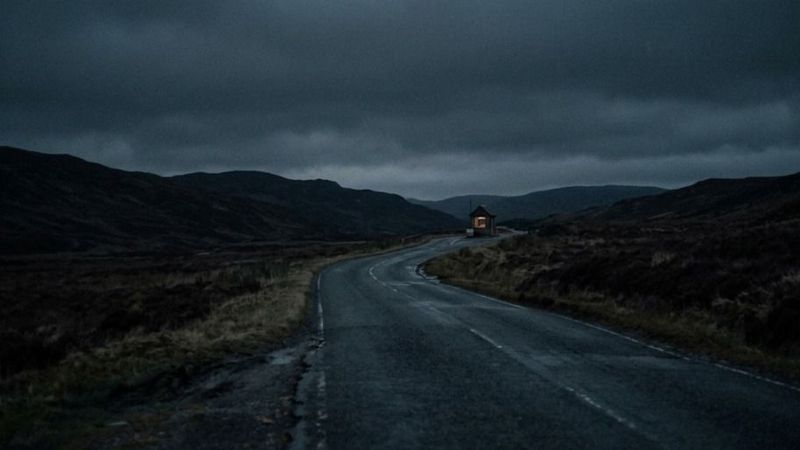 A winding road leading through a dark landscape toward a small illuminated building in the distance, symbolizing a long journey toward a goal.