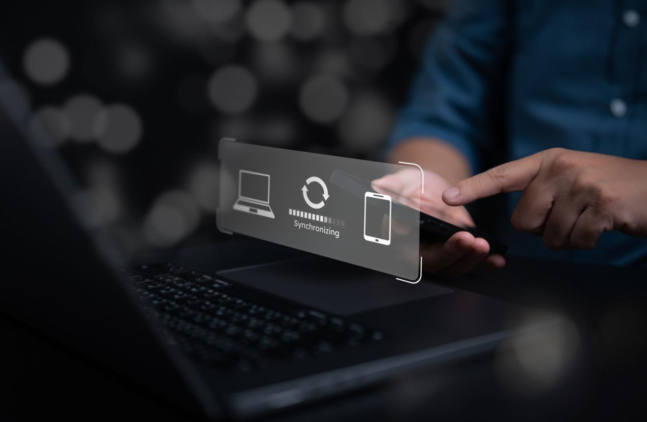 **Alt text:** Person using a smartphone near a laptop with a digital synchronization interface overlay, showing data syncing between mobile and desktop devices.