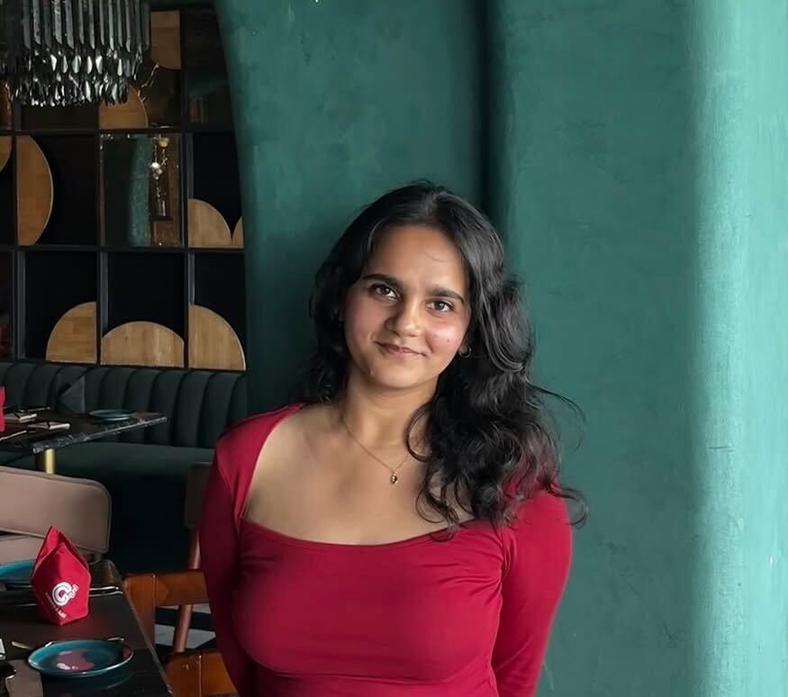 A woman wearing a red top stands indoors near a green pillar, smiling slightly, with a stylish restaurant setting and seating visible in the background.