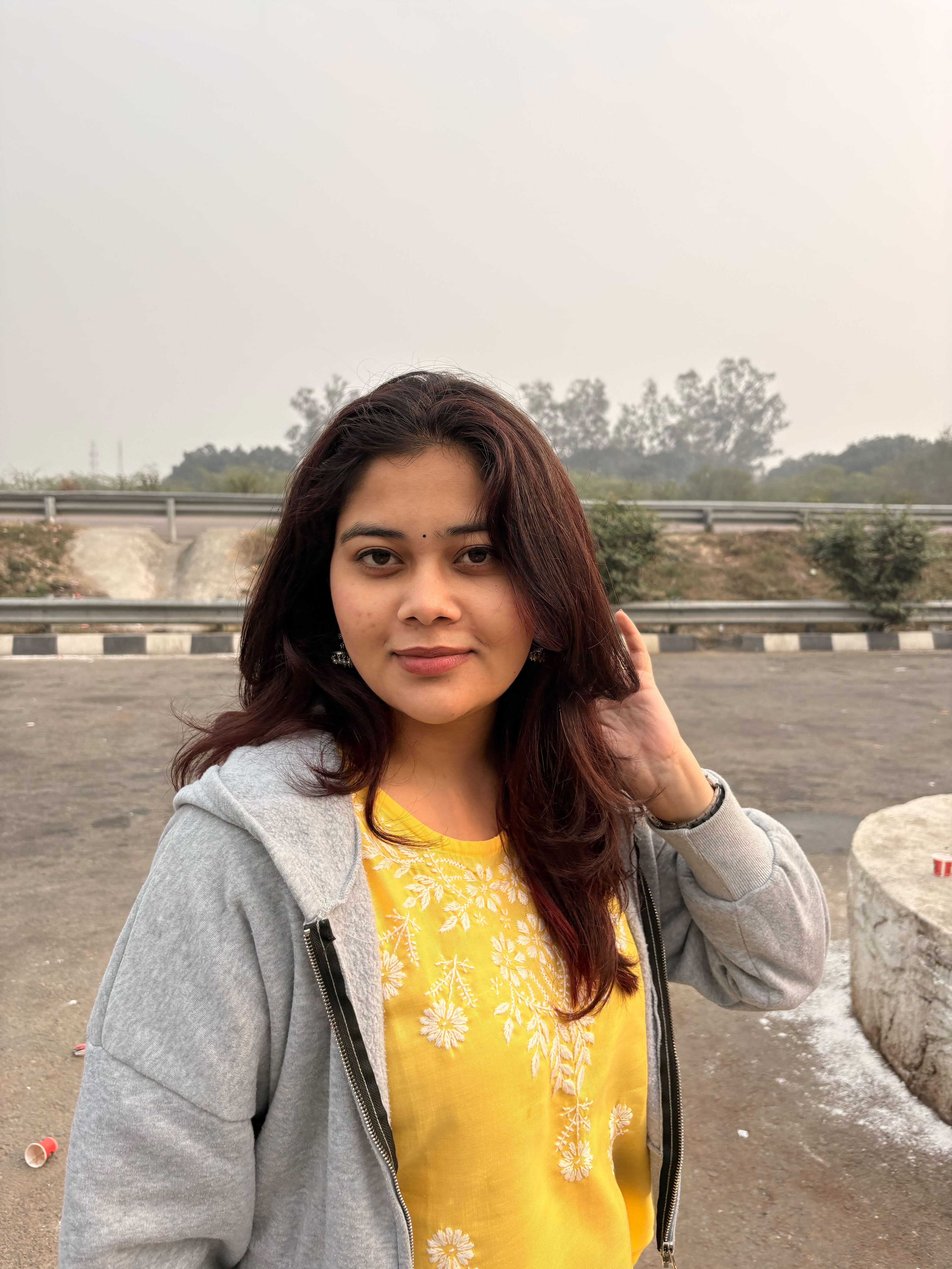 A woman wearing a yellow embroidered top and a gray hoodie stands outdoors near a roadside, gently touching her hair, with trees and a hazy sky in the background.